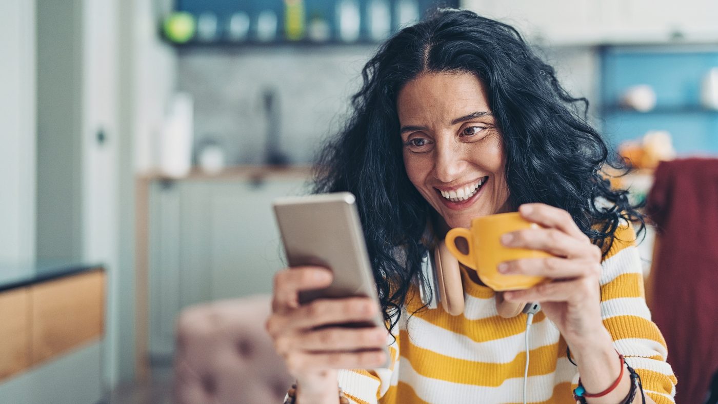 A woman smiling at her phone while holding a mug at home